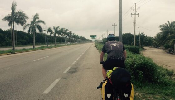 Photograph of Tom Passmore cycling along a Mexican road with a sigh to Tulum