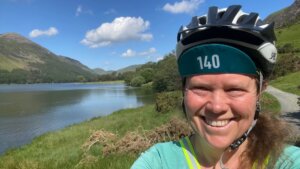 A selfie photograph of a smiling woman, Sophie Walker, wearing a cycling helmet with Buttermere lake in the background on a nice day.