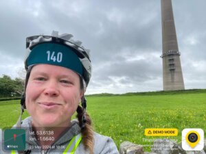 A selfie photograph of a woman, Sophie Walker, wearing a cycling helmet in front of a large stone tower called Emley Tower. 