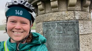 A photograph of a woman smiling into the camera wear a number 140 cap under the cycling helmet next to a metal sign saying Scar House Reservoir